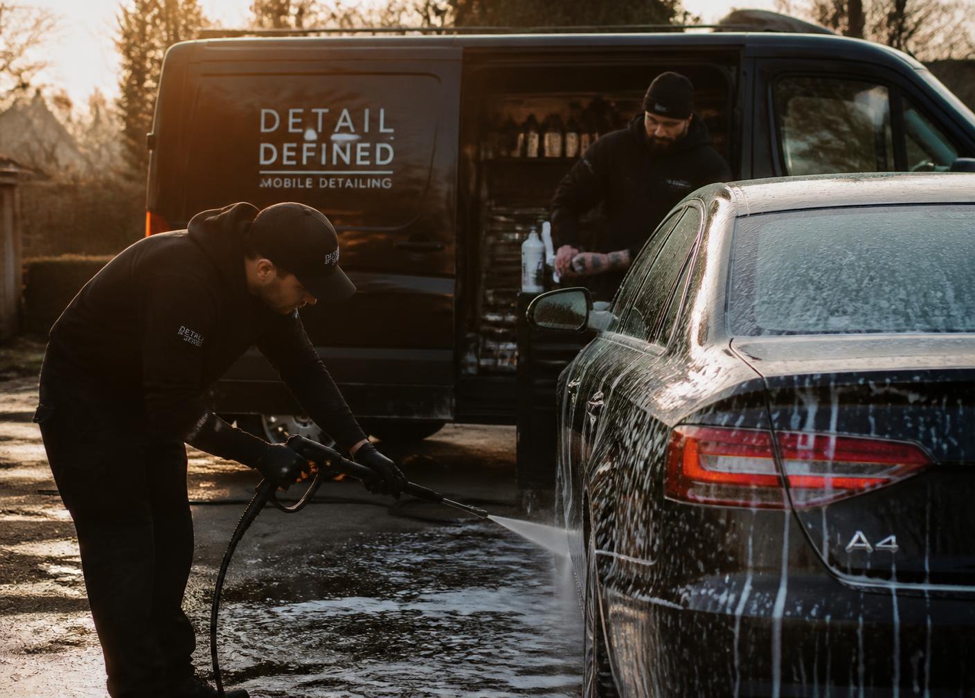 Winsome founders pressure washing a car beside a mobile detailing van at sunset